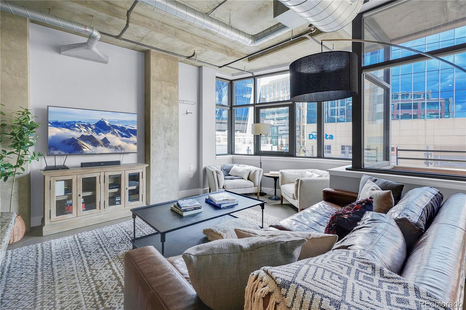 This is an interior shot of a modern living room featuring an exposed concrete ceiling with visible ductwork, large windows offering city views, and a comfortable seating arrangement. The room includes a large leather sofa with throw pillows and blankets, two armchairs, a coffee table with books, and a media console with a large television displaying a mountain scene. The overall aesthetic is industrial-chic with a touch of warmth and comfort.