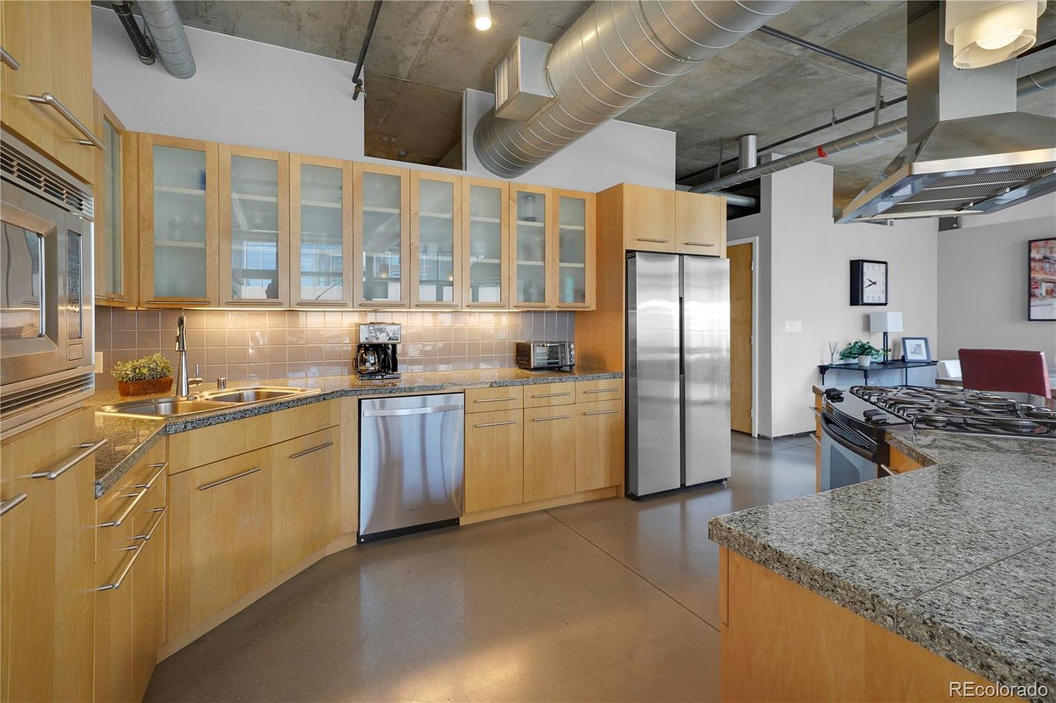 This is a well-lit kitchen with modern industrial design elements, featuring light wood cabinetry, stainless steel appliances, and granite countertops. The open layout includes a kitchen island with a cooktop and a stainless steel range hood. Exposed ductwork and a concrete ceiling add to the industrial aesthetic, while the polished concrete floors reflect the light, creating a spacious and airy feel.