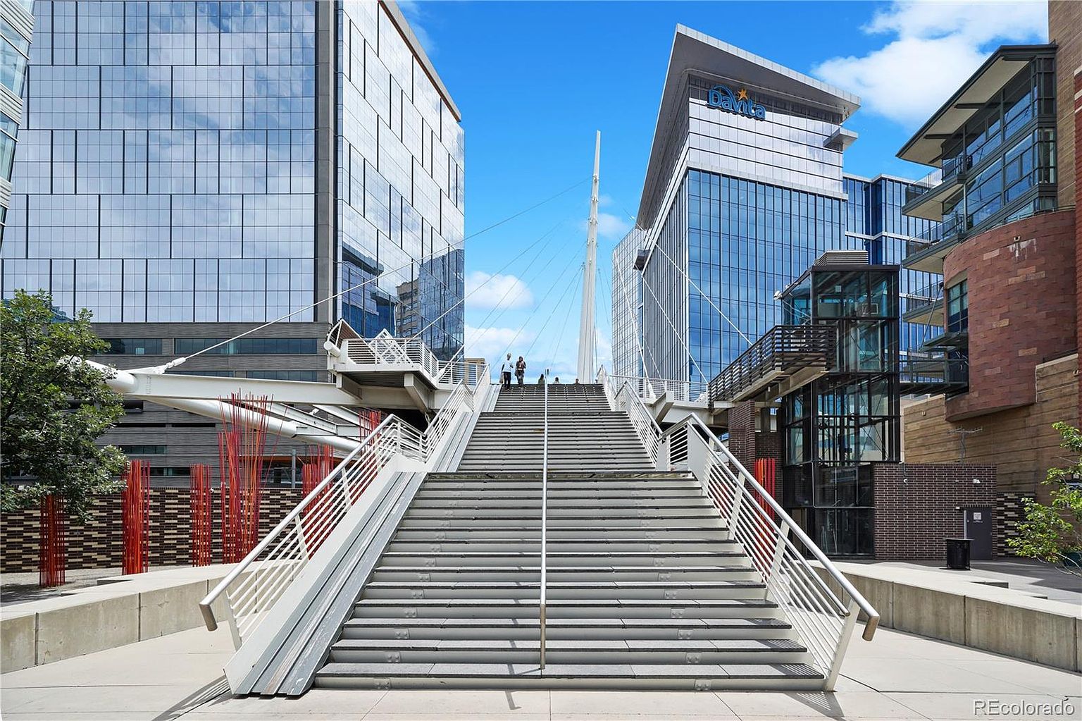This image showcases a modern architectural marvel, featuring a grand staircase leading up to a pedestrian bridge suspended between two contemporary buildings. The buildings boast sleek glass facades, reflecting the clear blue sky, while the white railings and support structure of the bridge create a striking contrast. The scene exudes a sense of urban sophistication and architectural innovation.