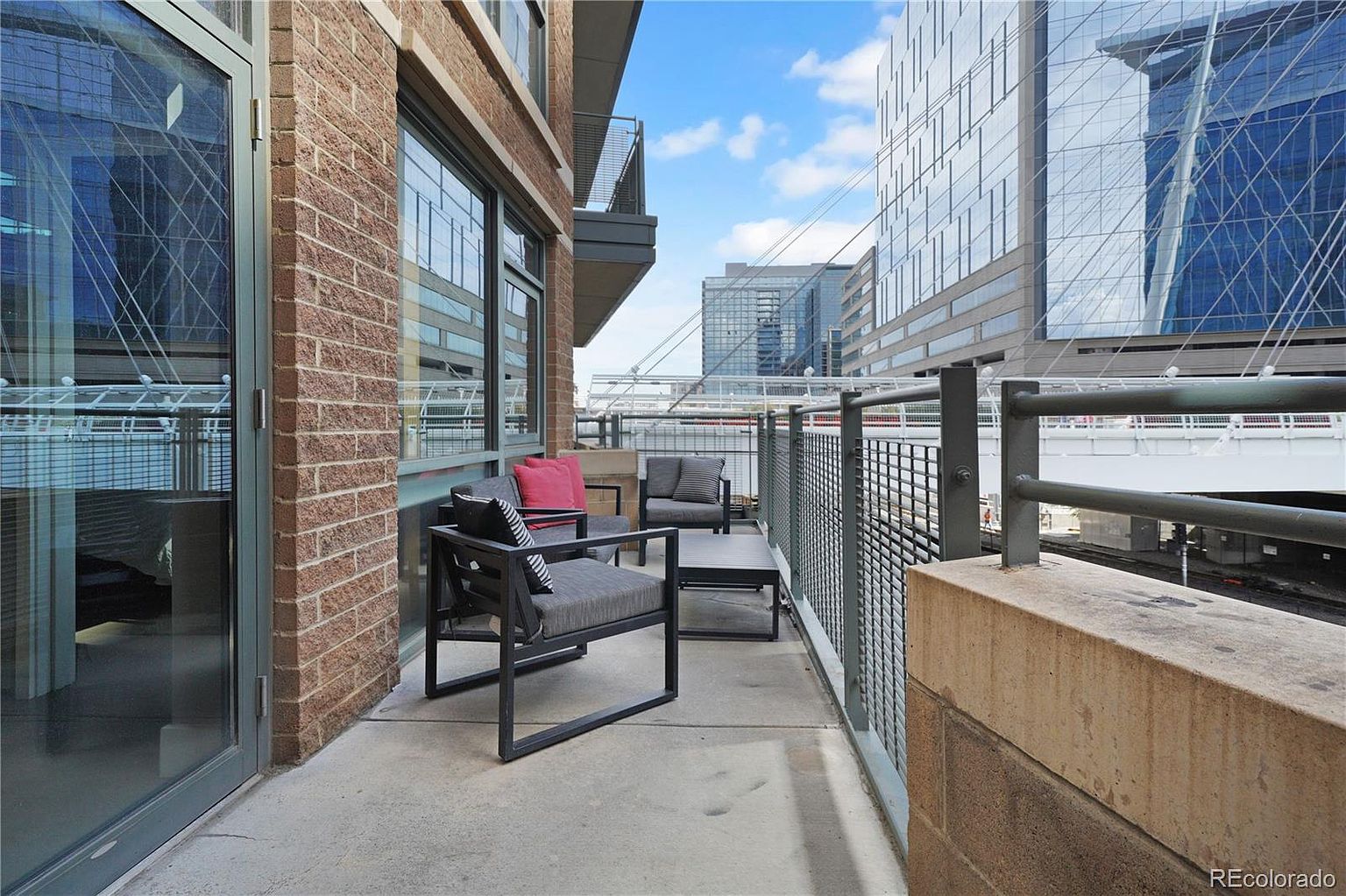 This image showcases a modern balcony with stylish outdoor furniture, including a lounge chair and a small table. The balcony features a brick facade and a metal railing, offering views of the surrounding cityscape and a nearby bridge. The space appears well-maintained and inviting, perfect for relaxing or entertaining.