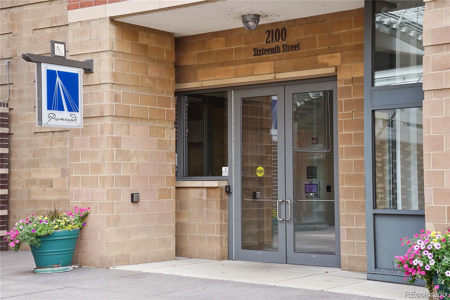 This image showcases the entryway of a building, featuring a brick facade and a glass double-door entrance. A sign with the building's name hangs on the left, and planters with colorful flowers add a welcoming touch. The address, "2100 Sixteenth Street," is displayed above the entrance, providing clear identification.
