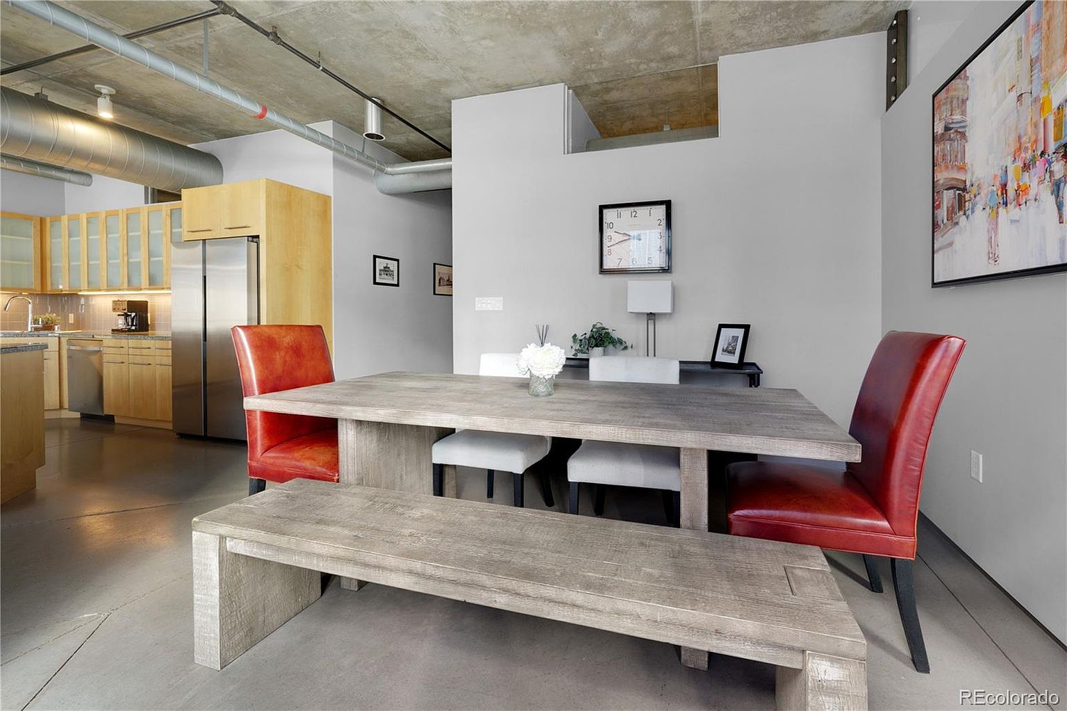 This interior shot showcases a modern dining area with a rustic touch. A large, weathered wooden table is surrounded by red leather chairs and a matching bench, complemented by white upholstered stools. The room features exposed ductwork and a concrete ceiling, adding an industrial feel, while a large painting and a clock on the wall contribute to the decor.