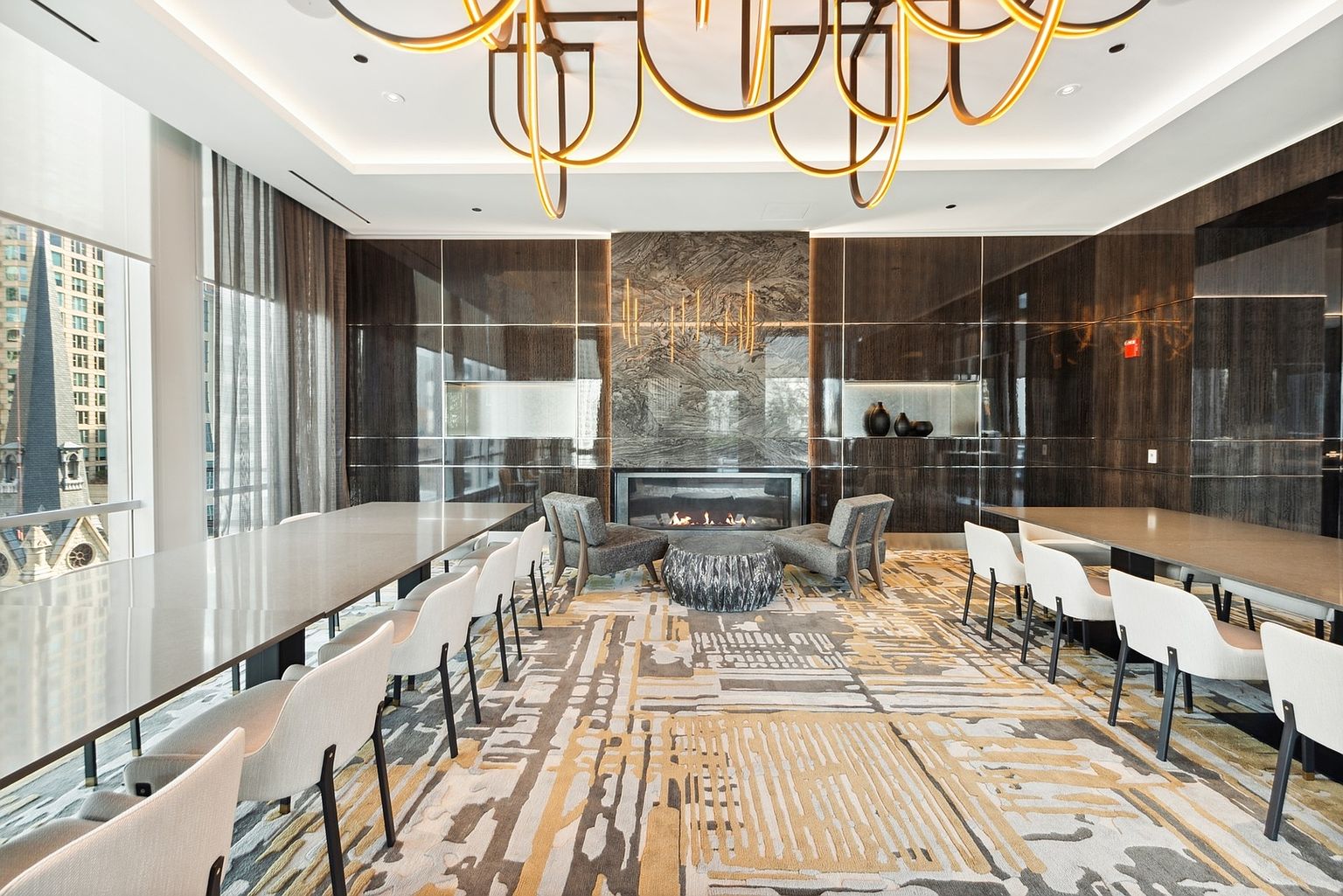 This is a stylish and modern living room featuring a fireplace as a focal point, surrounded by dark wood paneling and elegant lighting fixtures. The room is furnished with comfortable seating arrangements and long tables with chairs, creating a versatile space for relaxation and entertaining. A large window offers a view of the city skyline, enhancing the room's appeal.