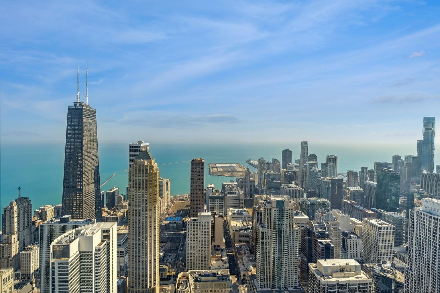 This aerial view showcases a sprawling cityscape with numerous skyscrapers and buildings, set against a backdrop of a large body of water and a clear blue sky. The iconic John Hancock Center stands prominently among the buildings. The image captures the scale and density of the urban environment, highlighting the city's architectural diversity and waterfront location.