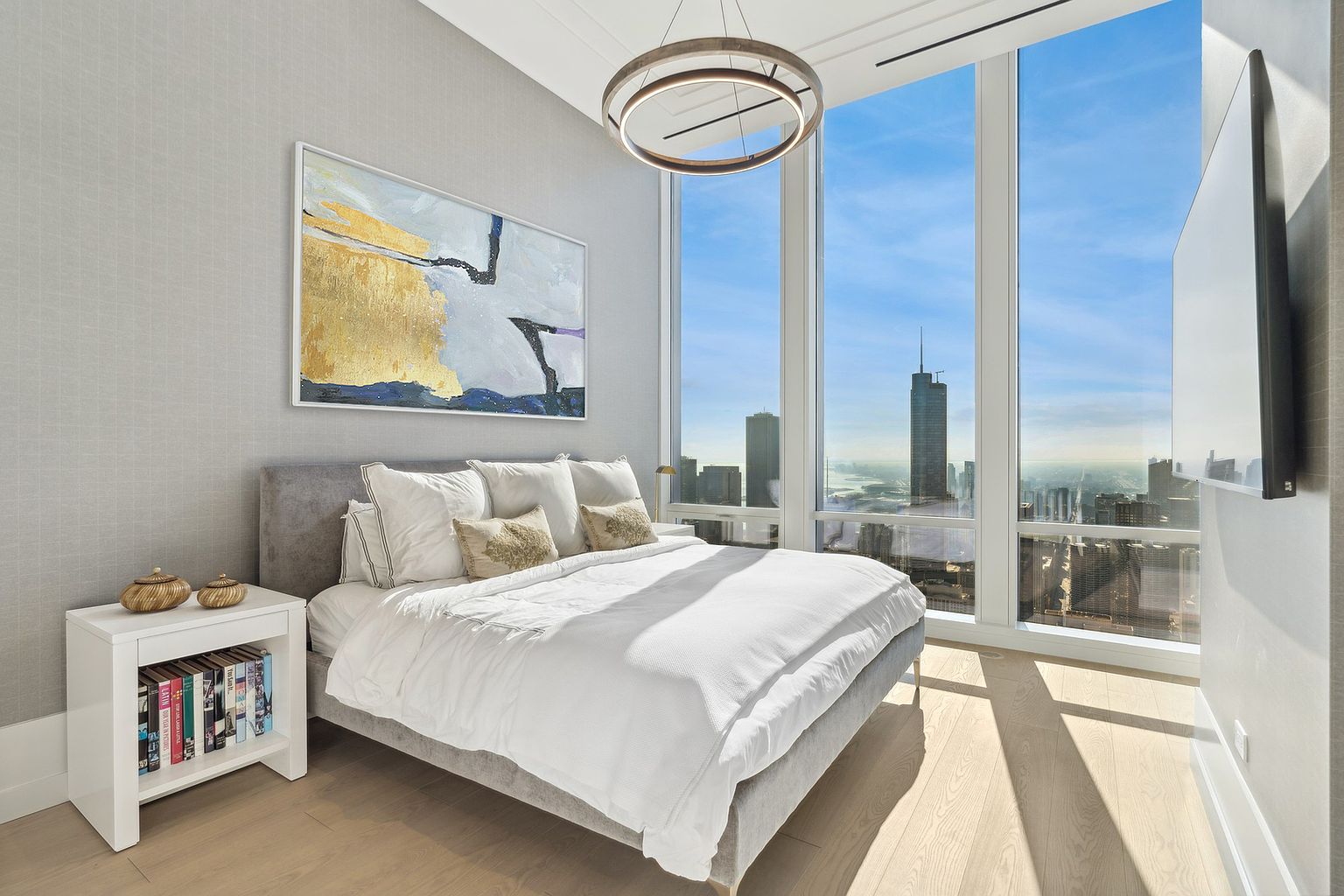 This is a bright and modern primary bedroom featuring floor-to-ceiling windows with a stunning city view. The room is decorated in neutral tones with a gray upholstered bed frame, white bedding, and a large abstract painting above the bed. A contemporary circular chandelier hangs from the ceiling, and a white nightstand with books sits beside the bed, adding to the room's sophisticated and comfortable ambiance.
