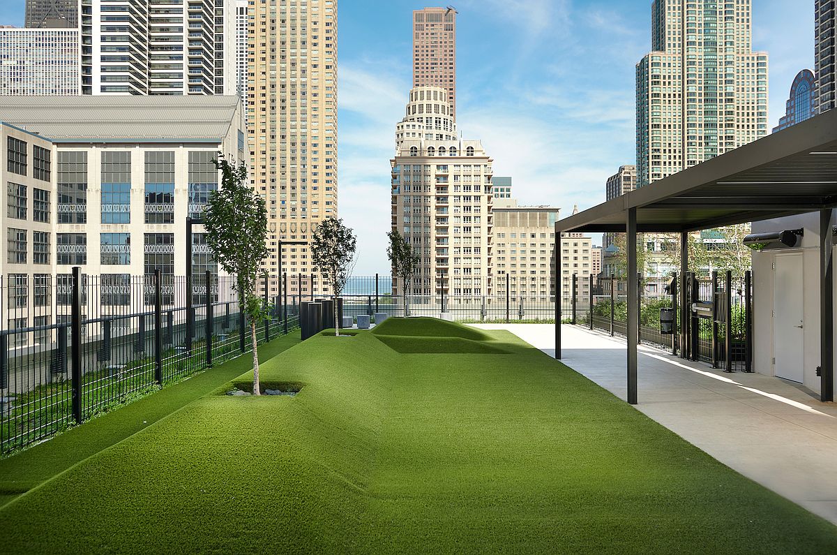 This image showcases a modern rooftop patio or deck area with artificial turf and landscaping, set against a backdrop of city skyscrapers. The space includes a covered area with a sleek, minimalist design, and a fence surrounds the perimeter. The overall impression is one of a stylish and functional outdoor space in an urban environment.