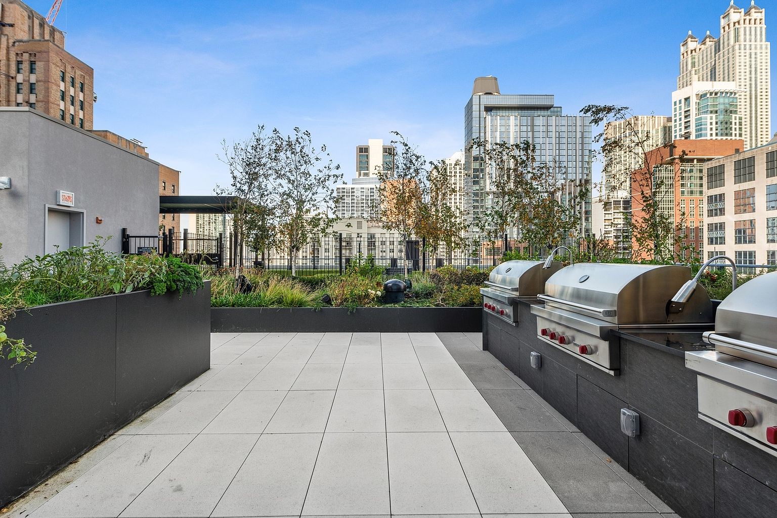 This image showcases a stylish rooftop patio or deck area, complete with built-in stainless steel grills and lush greenery in planters. The space features a clean, modern design with light-colored tile flooring and a backdrop of urban buildings under a clear blue sky. It presents an inviting outdoor space for entertaining and relaxation.