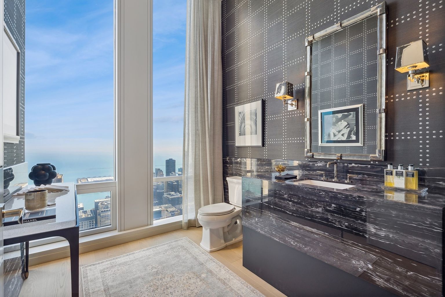 This is a stylish guest bathroom featuring a black marble countertop and backsplash, complemented by a patterned wallpaper. A large window offers a stunning city view, enhancing the luxurious feel of the space. The bathroom includes modern fixtures and a well-lit mirror, creating an elegant and functional design.