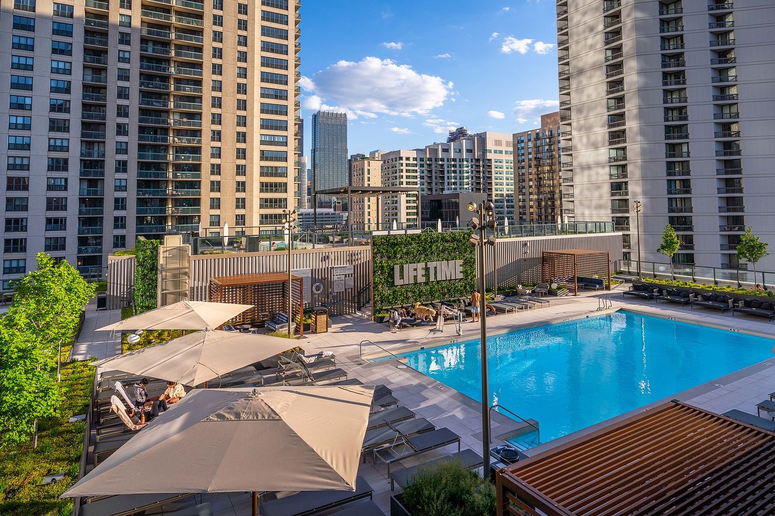 This image showcases a luxurious rooftop pool area surrounded by modern high-rise buildings. The pool is a vibrant blue, complemented by lounge chairs and umbrellas for relaxation. A green wall with the words "LIFE TIME" adds a touch of nature and branding to the space, creating an inviting and upscale amenity for residents.
