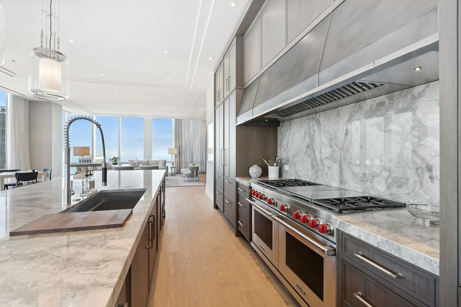 This is a high-end kitchen featuring a large island with a marble countertop and a stainless steel sink. The kitchen also includes a professional-grade Wolf range with a stainless steel hood and marble backsplash. The space is open to a living area with large windows offering a view of the city and the ocean, creating a luxurious and modern atmosphere.