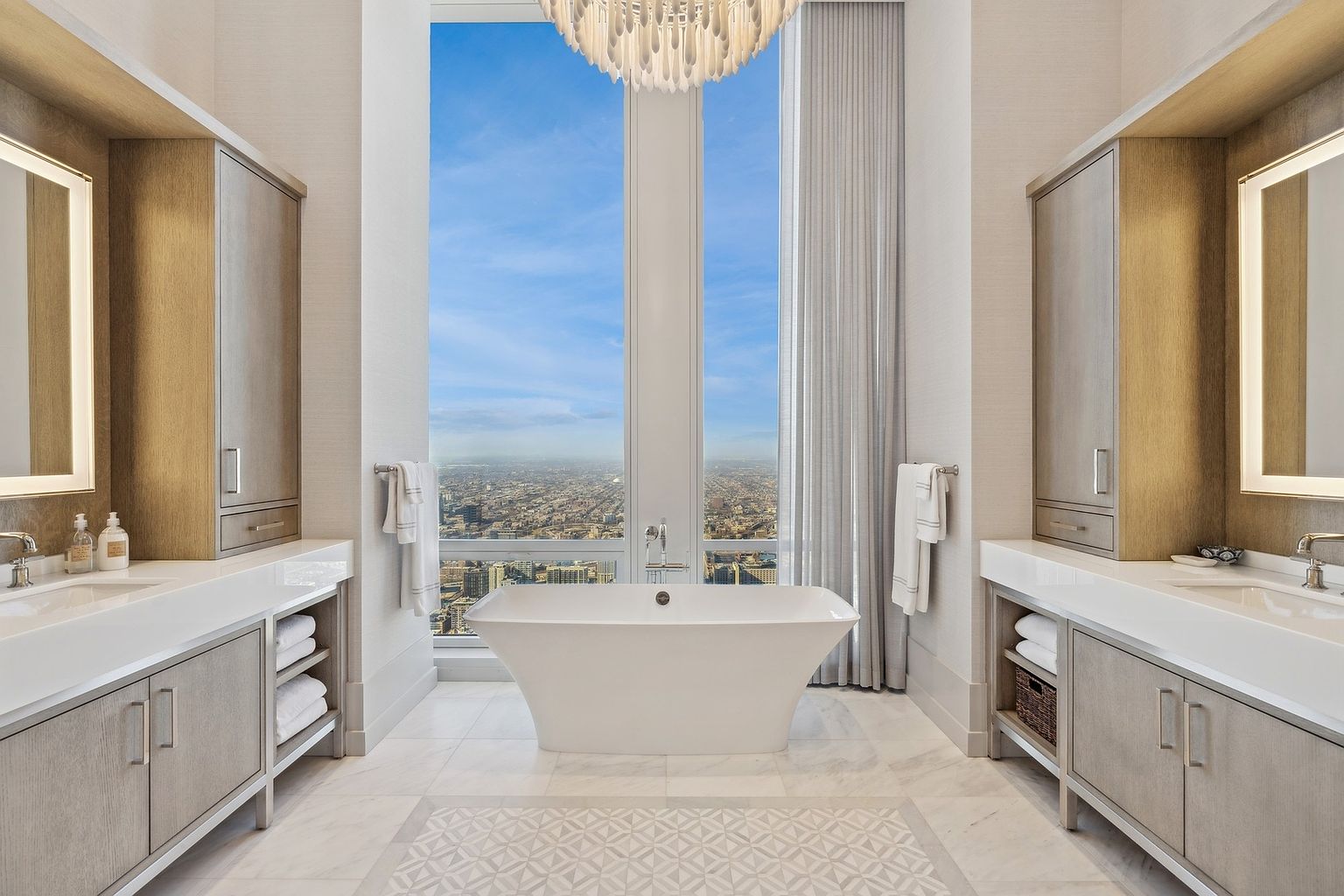 This is a luxurious primary bathroom featuring dual vanities with light wood cabinetry and white countertops, flanking a freestanding white bathtub positioned in front of a large window with a city view. The room is illuminated by a modern chandelier and recessed lighting, creating a bright and elegant atmosphere. The floor is tiled with a decorative pattern, adding a touch of sophistication.
