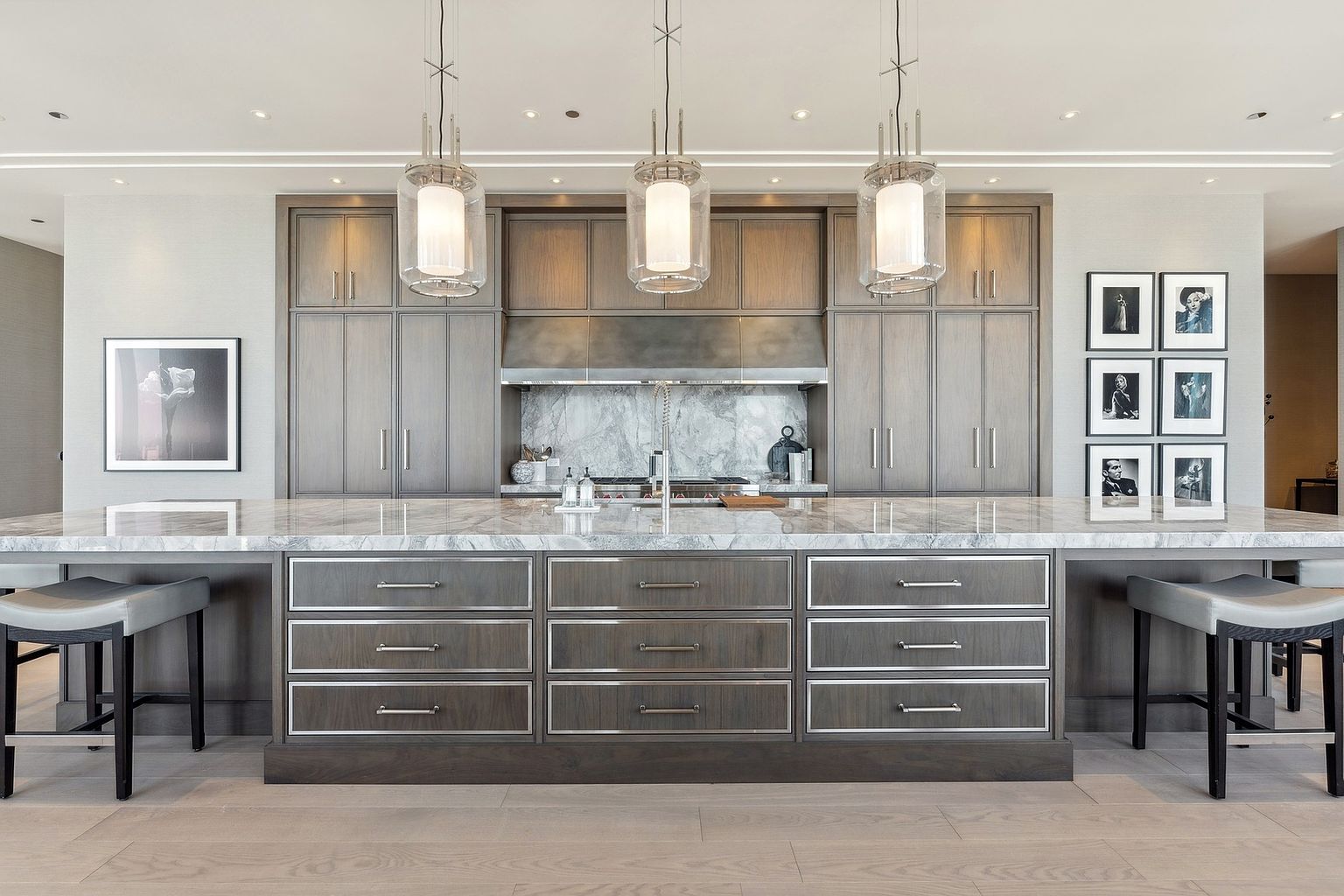 This is a well-lit kitchen featuring a large island with a marble countertop and dark wood cabinetry with silver hardware. Three pendant lights hang above the island, and the kitchen is equipped with stainless steel appliances and a marble backsplash. The perspective is at eye level, showcasing the island and cabinetry in detail.