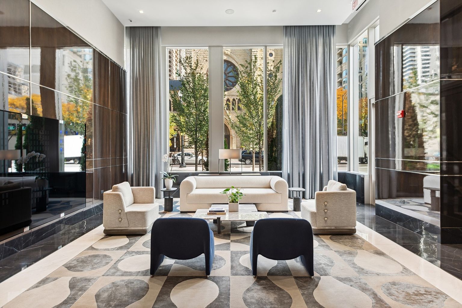 This is a well-lit and stylish living room featuring a neutral color palette with pops of navy blue. The room is furnished with a cream-colored sofa, two matching armchairs, and two navy blue accent chairs. A marble coffee table sits in the center of the room, and large windows offer a view of a building across the street, creating an inviting and sophisticated atmosphere.