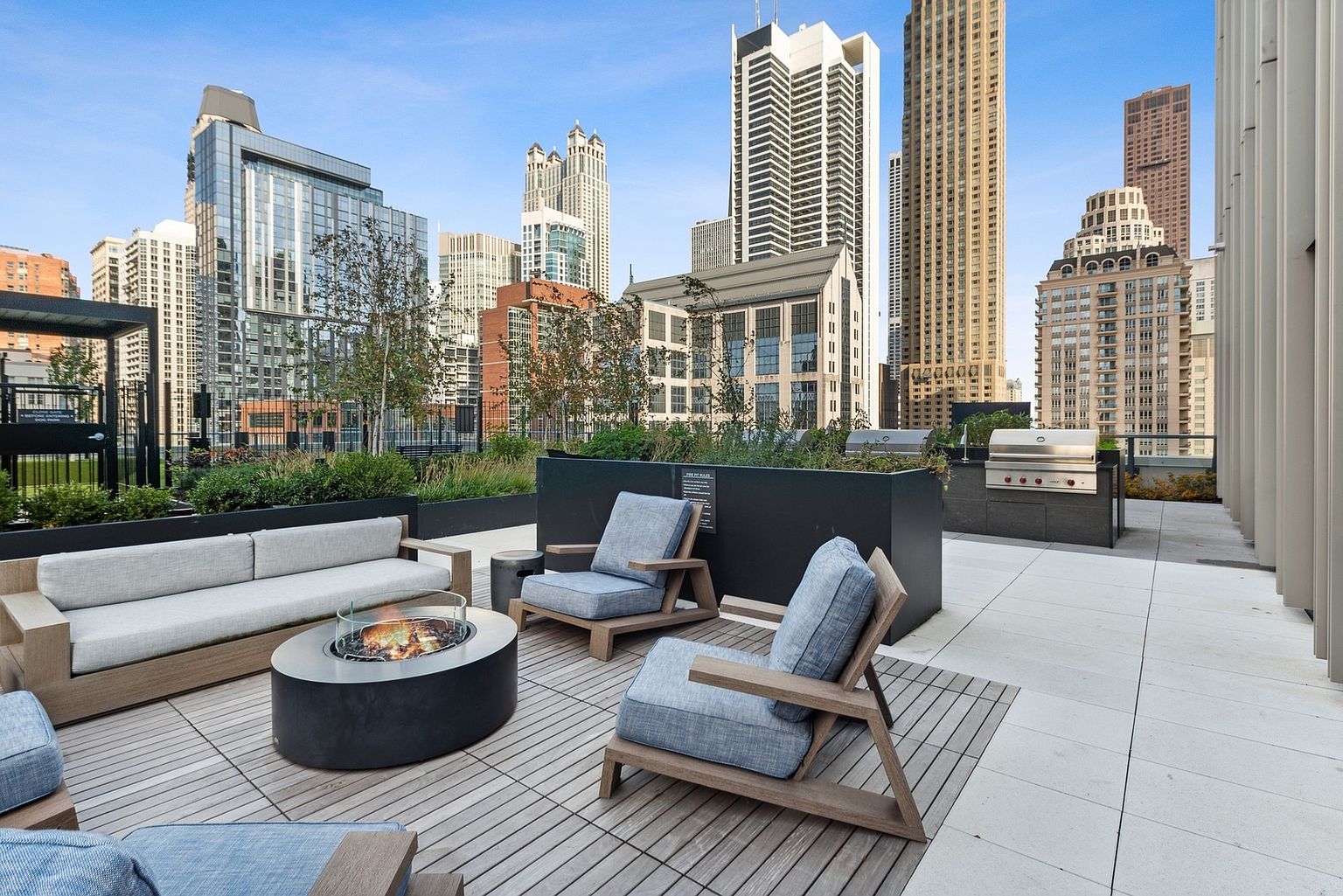 This image showcases a luxurious rooftop patio with stunning city views. The space is furnished with comfortable seating around a modern fire pit, complemented by a built-in grill area and lush planters. The overall impression is one of upscale urban living and sophisticated outdoor entertaining.