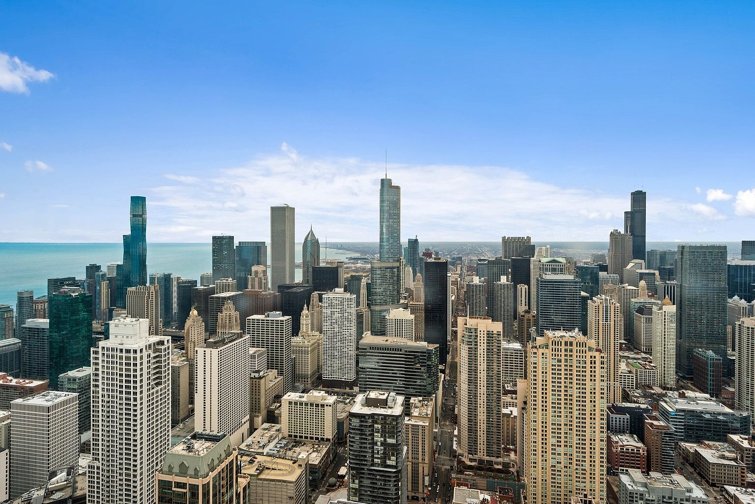 This aerial view showcases a dense cityscape with numerous skyscrapers under a clear blue sky. The buildings vary in height and architectural style, creating a dynamic urban landscape. A body of water is visible in the distance, adding depth to the scene and suggesting a waterfront location.