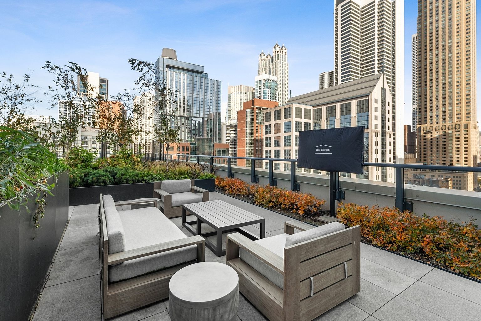 This image showcases a luxurious rooftop patio or balcony with comfortable outdoor furniture, including a sofa, chairs, and a coffee table. The space is surrounded by lush greenery and offers stunning views of the city skyline with modern high-rise buildings. A covered television is also present, suggesting a space for outdoor entertainment.