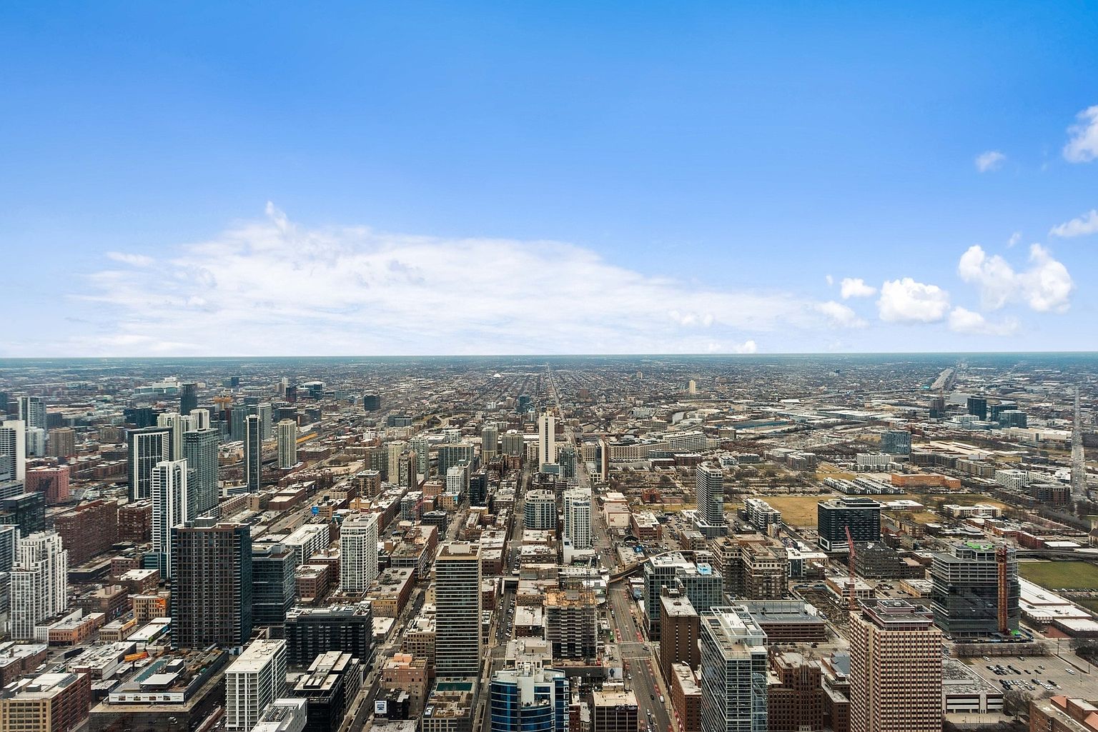This aerial view showcases a sprawling cityscape under a bright blue sky with scattered clouds. The urban landscape is densely packed with buildings of varying heights and architectural styles, intersected by numerous streets and avenues. The perspective provides a comprehensive overview of the city's scale and layout, emphasizing its vibrant and bustling atmosphere.