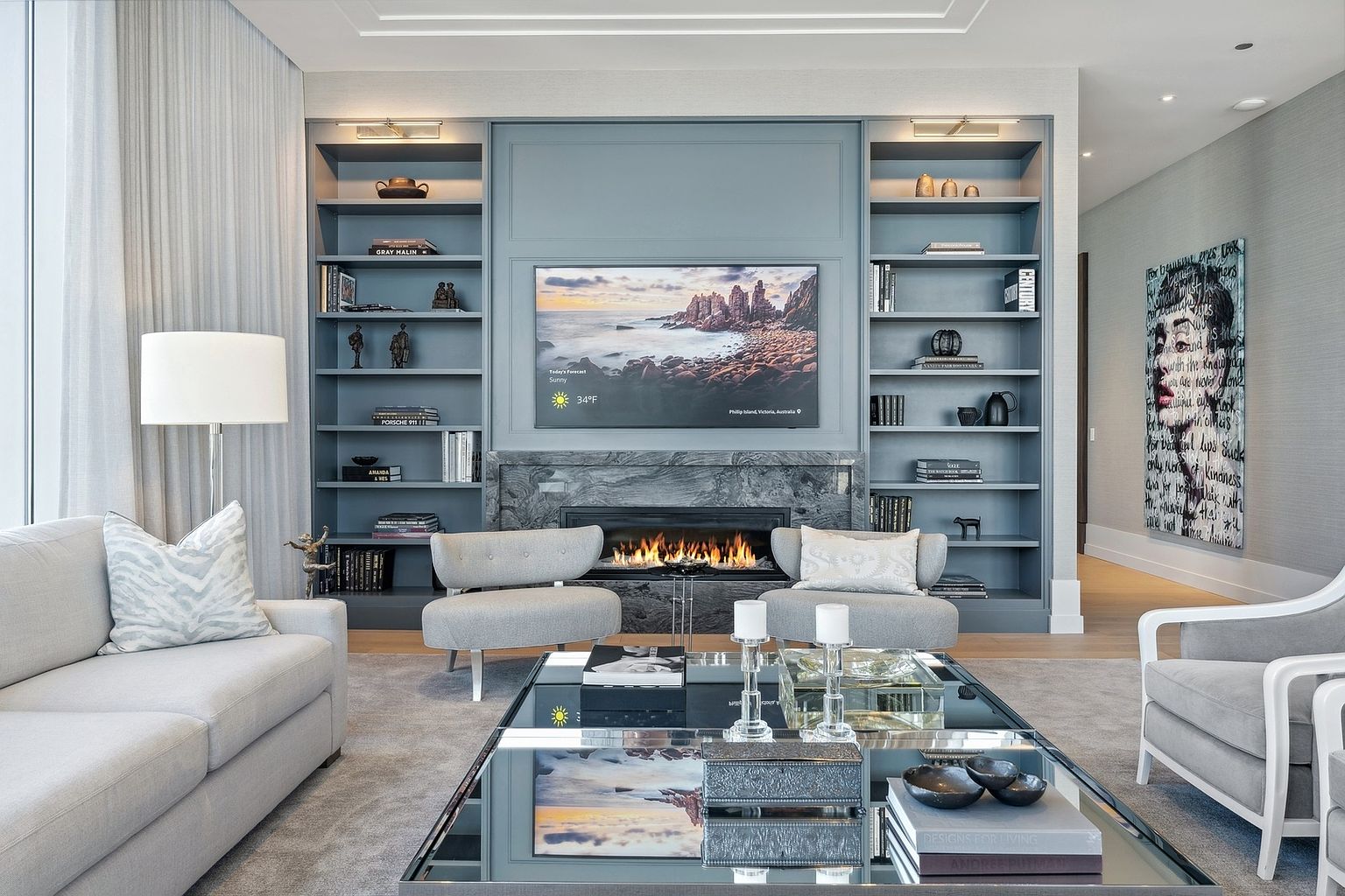 This is a beautifully designed living room featuring a built-in shelving unit with a fireplace and a television. The room is decorated in a modern style with neutral colors and elegant furnishings, including a comfortable sofa, armchairs, and a mirrored coffee table. The artwork and accessories add a touch of sophistication to the space.