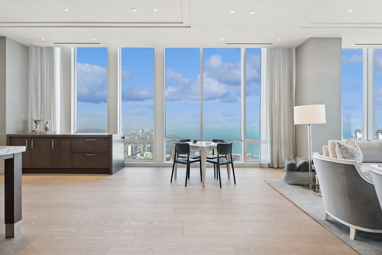 This is a bright and airy living room with floor-to-ceiling windows offering panoramic city and water views. The space features a modern aesthetic with light wood flooring, a gray sofa, and a small dining table with chairs. A built-in cabinet provides storage, and sheer curtains soften the natural light, creating a serene and luxurious atmosphere.