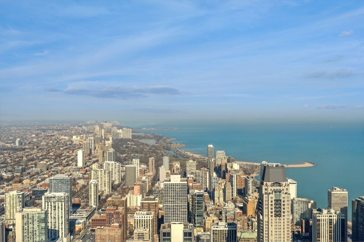 This aerial view showcases a dense urban landscape with numerous high-rise buildings, extending towards a large body of water under a clear blue sky. The city's architecture is a mix of modern and classic styles, creating a dynamic skyline. The image emphasizes the scale and location of the city, highlighting its proximity to the waterfront.