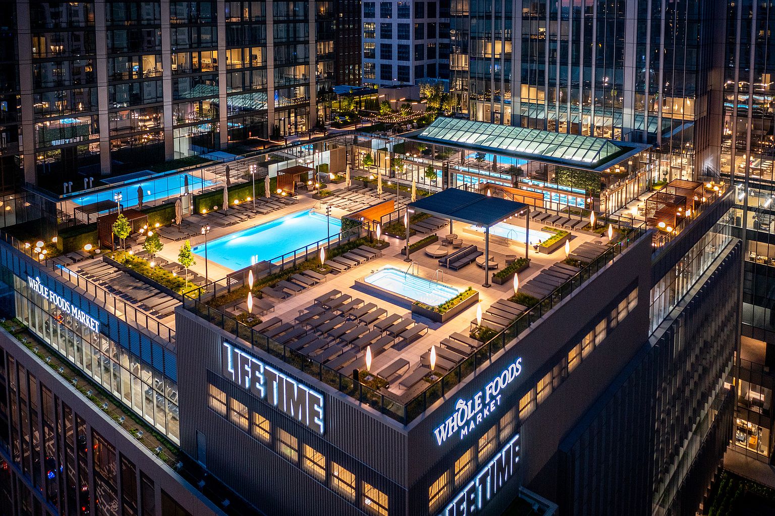 This aerial view showcases a luxurious rooftop amenity space, featuring multiple swimming pools, lounge areas with modern furniture, and lush landscaping. The space is integrated with the building's architecture, which includes retail spaces like Whole Foods Market and Life Time fitness center, creating a vibrant urban oasis. The overall impression is one of upscale living and sophisticated design.