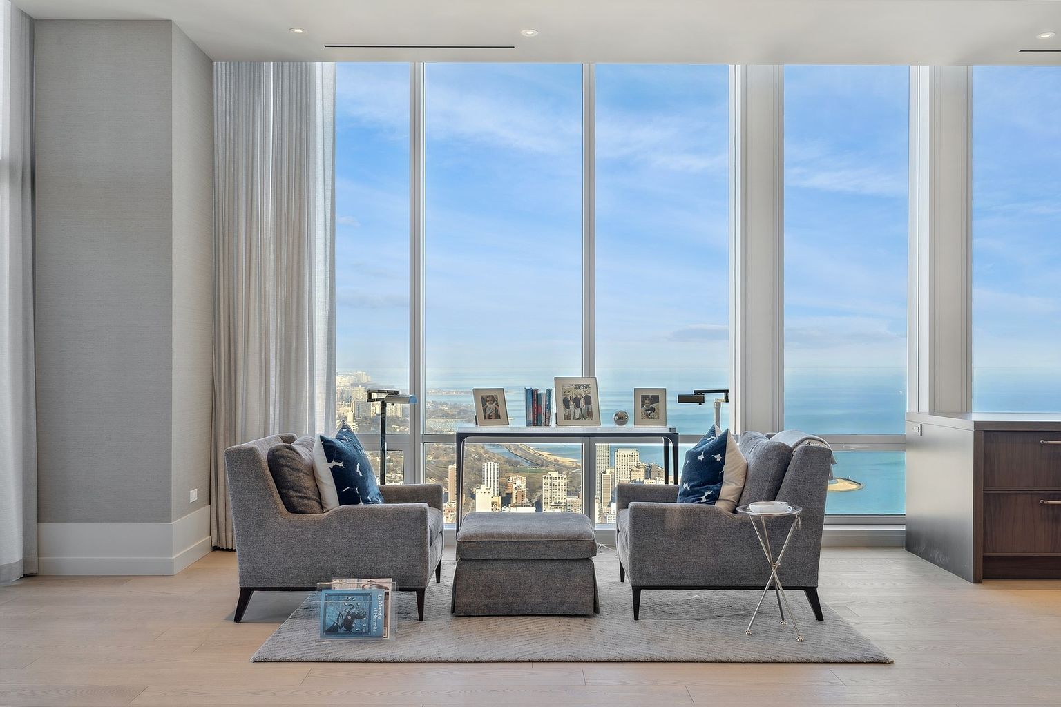 This is a bright and airy living room featuring floor-to-ceiling windows with stunning city and lake views. The room is furnished with two gray armchairs, a matching ottoman, and a glass-topped side table, all arranged on a light gray rug. The neutral color palette and minimalist decor create a sophisticated and relaxing atmosphere.