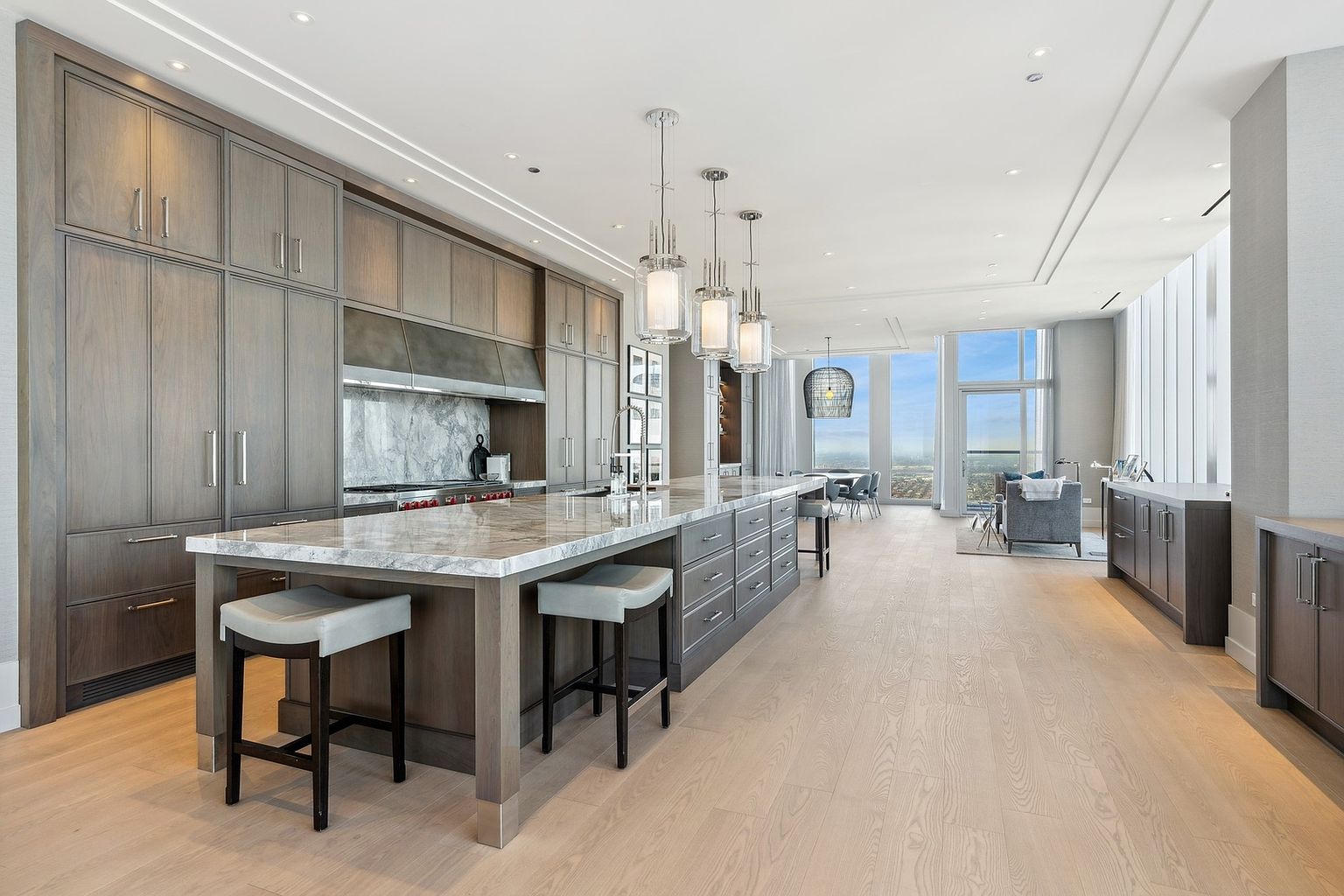 This is a spacious, modern kitchen featuring custom gray cabinetry, a large marble island with seating, and stainless steel appliances. The open floor plan seamlessly connects the kitchen to the dining and living areas, offering expansive views through large windows. The overall design is luxurious and contemporary, perfect for entertaining.