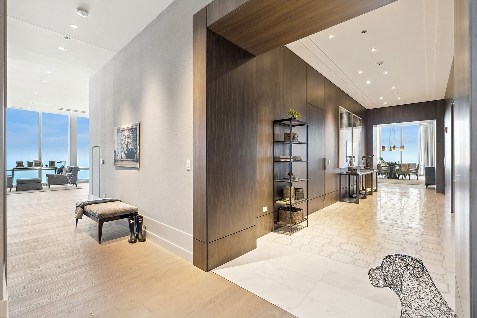 This interior shot showcases a luxurious hallway with a blend of light wood flooring and patterned tile. The hallway features a dark wood accent wall with built-in storage and a decorative shelving unit. Large windows at the end of the hallway offer a glimpse of a stunning view, enhancing the sense of spaciousness and elegance.