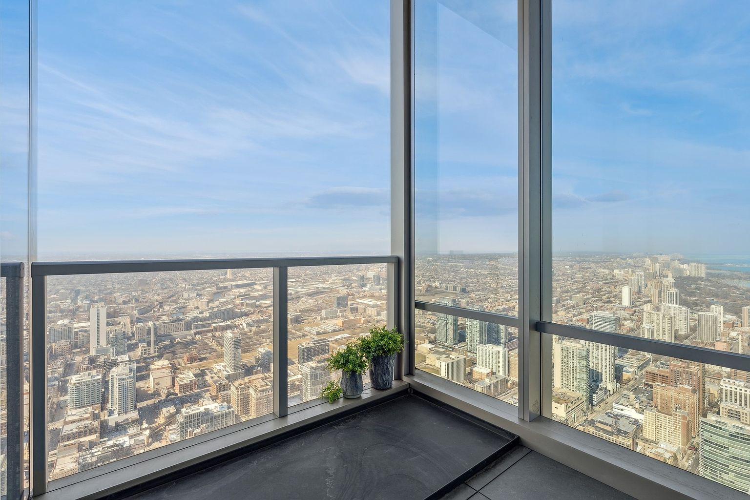 This image showcases a modern balcony with stunning panoramic city views. The balcony features a sleek, dark-toned floor and a glass railing that provides an unobstructed view of the cityscape. Two potted plants add a touch of greenery to the space, enhancing the overall aesthetic.