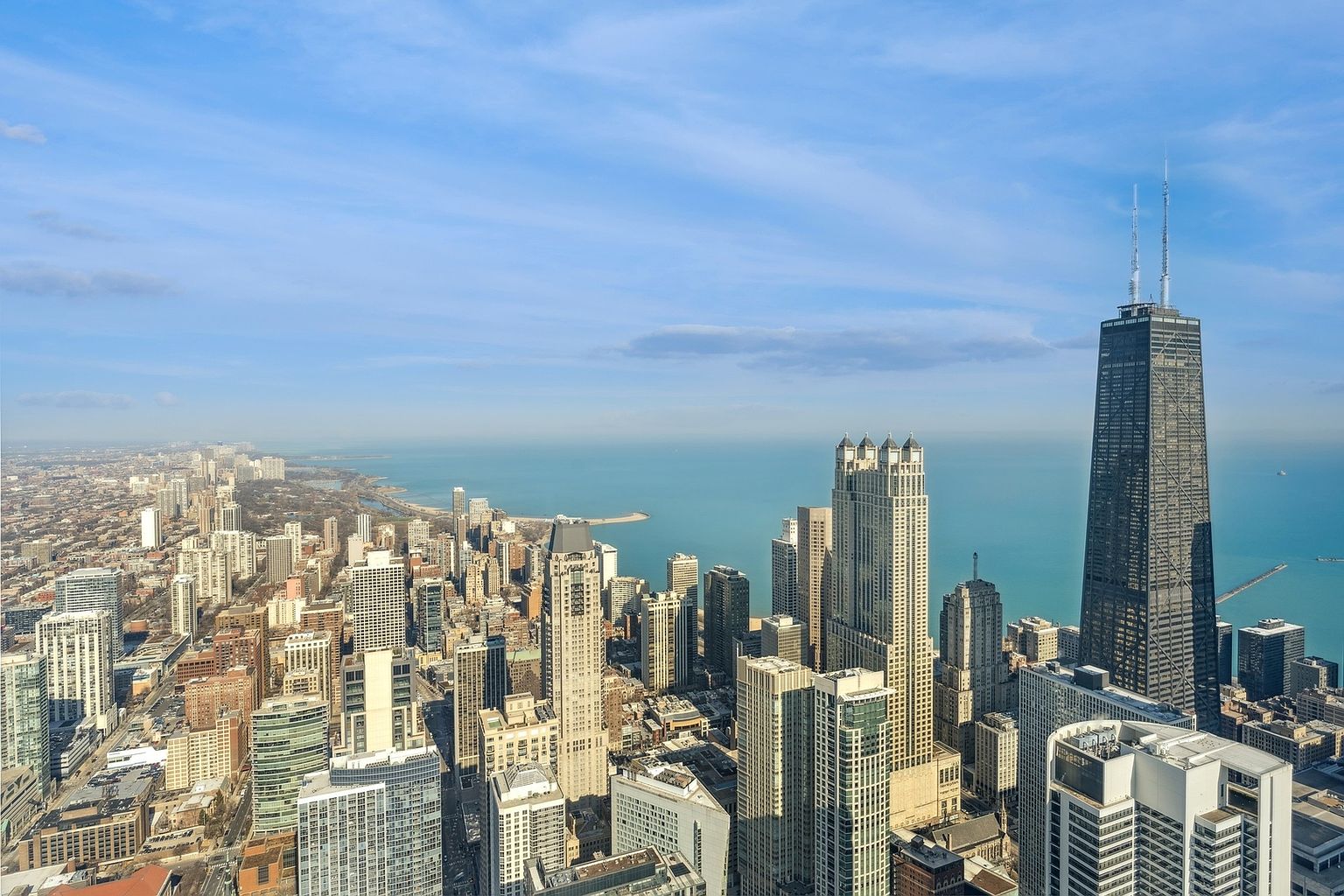 This aerial view showcases the impressive Chicago skyline, dominated by iconic skyscrapers and the vast expanse of Lake Michigan. The cityscape is a mix of architectural styles, with modern high-rises and classic buildings creating a dynamic visual. The clear blue sky and shimmering lake enhance the overall grandeur and appeal of this urban landscape.