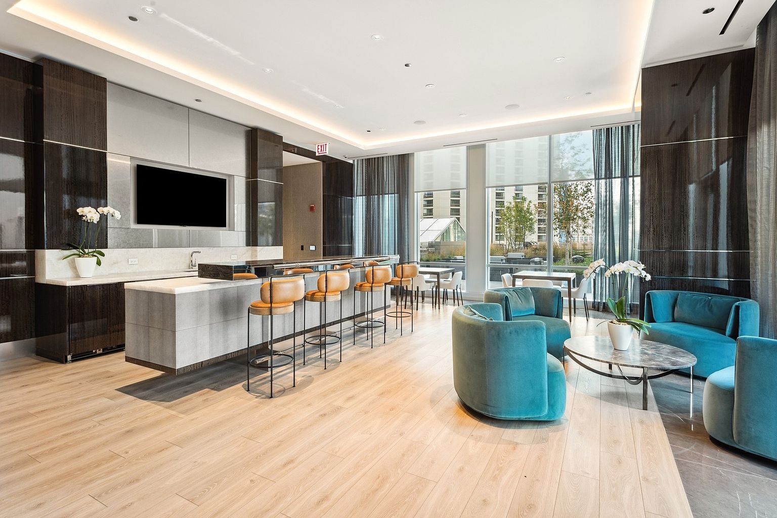 This is a well-appointed community amenity space featuring a modern kitchen area with bar seating and a comfortable lounge area with plush teal seating. Large windows provide ample natural light and views of the outdoors, enhancing the inviting atmosphere. The space is designed for relaxation and socializing, making it an attractive feature for potential residents.