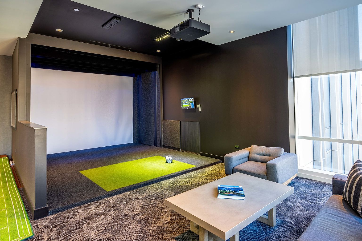 This is an interior shot of a home golf simulator room. The room features a large projection screen, a projector mounted on the ceiling, a putting green, and a seating area with a couch and armchair. The room is well-lit and appears to be designed for entertainment and recreation.