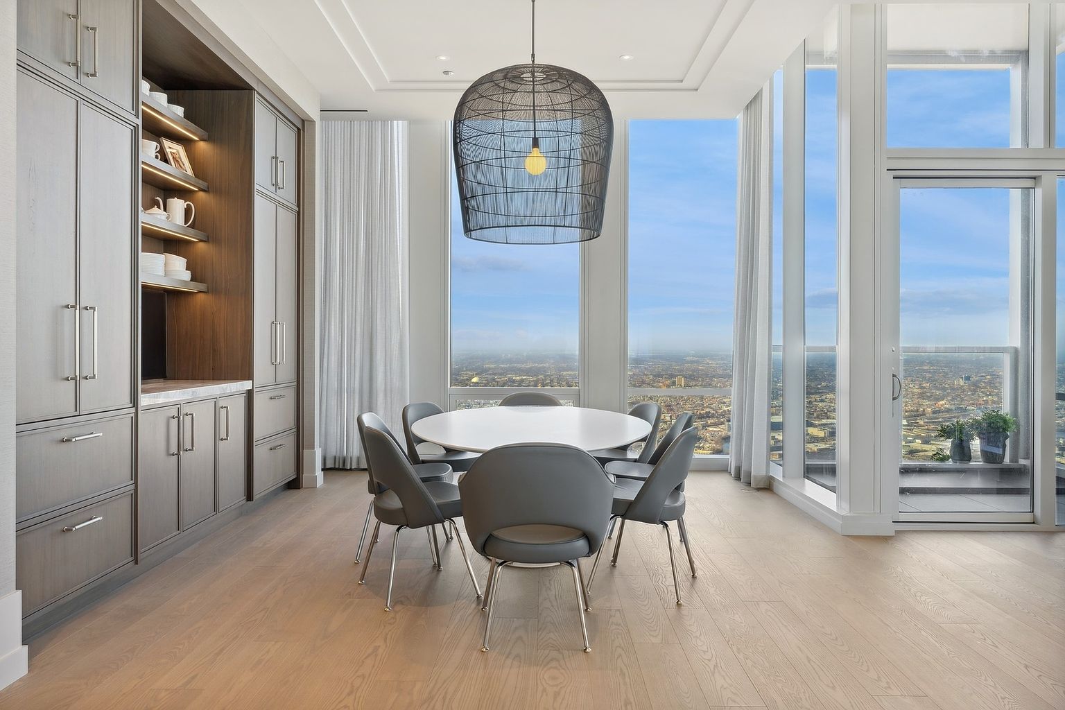 This is a bright and modern dining room featuring a round white table surrounded by six gray chairs with chrome legs. A large, sculptural pendant light hangs above the table, and floor-to-ceiling windows offer a panoramic city view. Built-in cabinetry provides ample storage, and the light wood flooring adds warmth to the space.