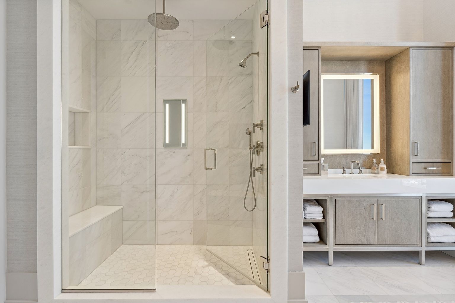 This is a luxurious primary bathroom featuring a glass-enclosed shower with marble tiling and a built-in bench. The shower includes a rainfall showerhead and a handheld sprayer. Adjacent to the shower is a vanity area with a large, illuminated mirror and ample storage, creating a spa-like atmosphere.