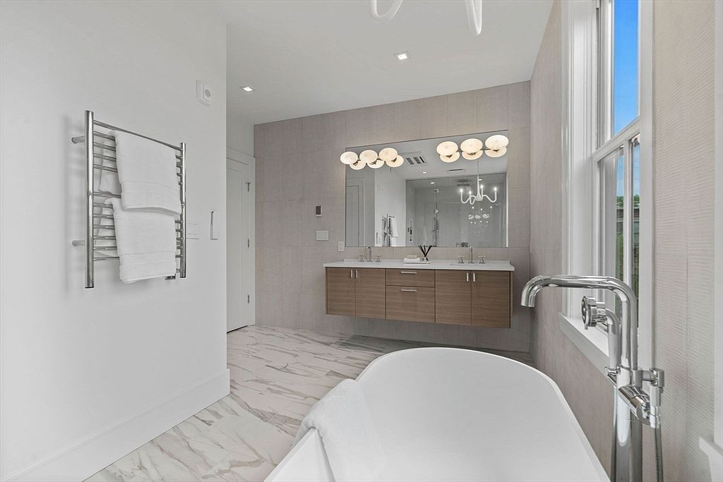 This is a bright and modern primary bathroom featuring a freestanding bathtub, a double vanity with a wood-grain cabinet, and a large mirror illuminated by stylish lighting. The floor is covered in marble-like tiles, and a heated towel rack is mounted on the wall. The overall impression is luxurious and spa-like.