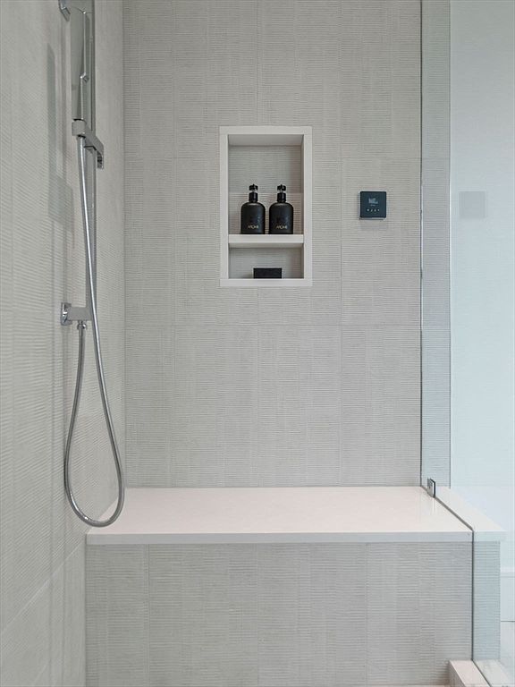 This is a detailed shot of a modern primary bathroom shower. The shower features light gray textured tiles, a built-in shelf with toiletries, a shower bench, and a sleek shower head. The overall impression is clean, minimalist, and luxurious.