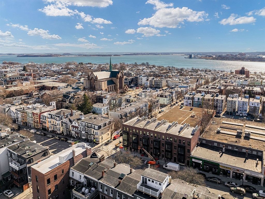 This aerial shot showcases a dense urban neighborhood with a mix of residential and commercial buildings. A prominent church with a steeple stands out amidst the rooftops. The scene extends to a waterfront with a visible coastline and a clear blue sky dotted with clouds, creating a vibrant and appealing overview of the area.
