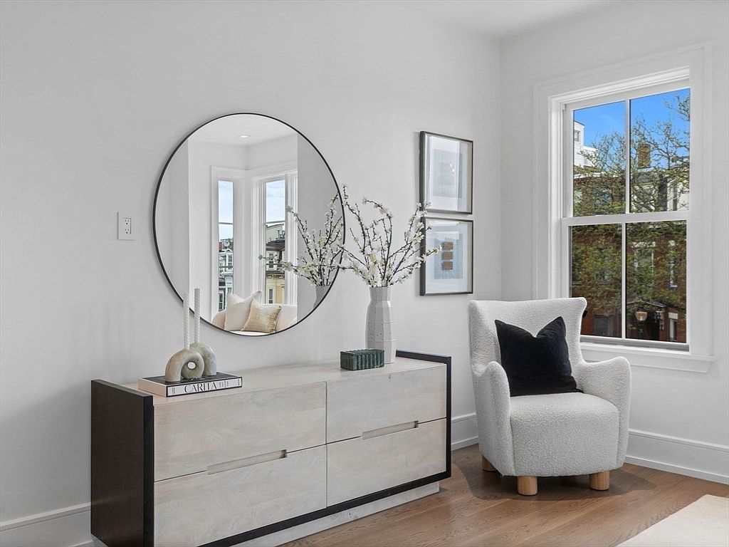 This interior shot showcases a stylish hallway or entryway featuring a modern dresser with a round mirror above it. The space is decorated with vases, books, and framed artwork, complemented by a comfortable armchair near a window that provides natural light. The overall aesthetic is clean, bright, and inviting, suggesting a well-designed and cared-for home.