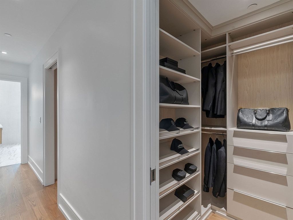 This image showcases a well-organized walk-in closet with custom shelving and drawers. The closet features a combination of open shelving for items like bags and hats, and hanging rods for clothing. The color palette is neutral, with white shelving and light wood accents, creating a clean and luxurious feel.