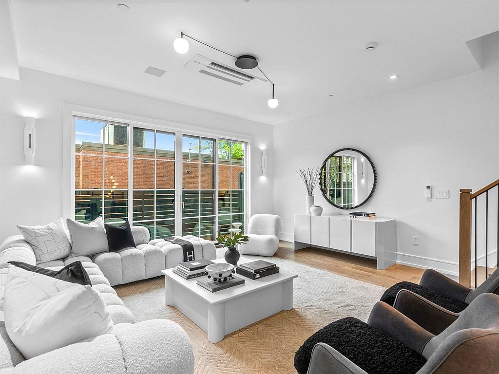 This is a bright and modern living room featuring a large window that provides ample natural light. The room is furnished with a white, textured sectional sofa, a white coffee table, and several armchairs. A round mirror hangs above a white console table, and the overall aesthetic is clean, minimalist, and inviting.