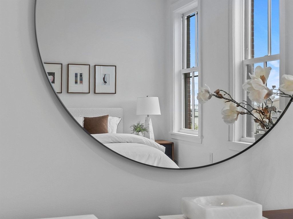 The image showcases a bedroom reflected in a round mirror, creating a sense of depth and space. The bedroom features a bed with white linens and a brown pillow, complemented by framed artwork on the wall. Natural light streams in through the windows, enhancing the bright and airy atmosphere of the room.