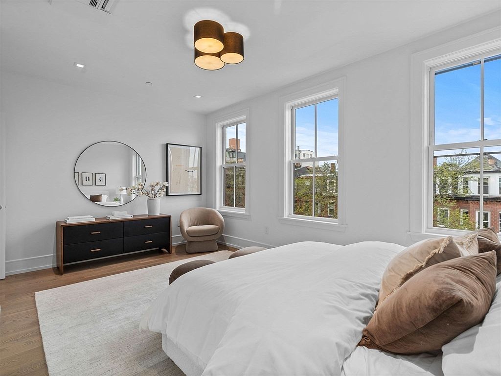 This is a bright and airy primary bedroom featuring a large bed with white linens and brown pillows, a dark wood dresser with a round mirror above, and a comfortable armchair. Two large windows provide ample natural light and views of the neighborhood. The room is decorated in a minimalist style with white walls and light wood floors, creating a serene and inviting atmosphere.