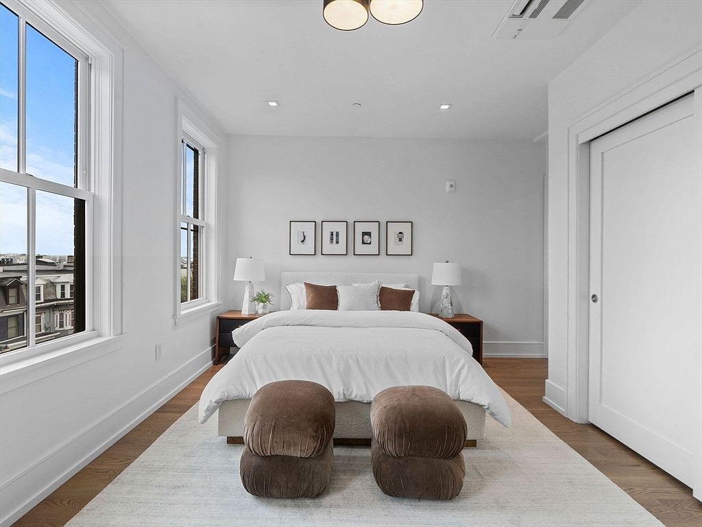 This is a bright and airy primary bedroom featuring hardwood floors, white walls, and large windows that offer a city view. The room is furnished with a bed, two nightstands, lamps, and two brown ottomans. Four framed pictures hang above the bed, adding a touch of elegance.