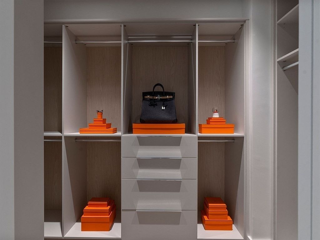 The image showcases a well-organized walk-in closet with a neutral color palette. The closet features built-in shelving, drawers, and hanging rods, providing ample storage space. Orange boxes and a black handbag add a touch of color and luxury, suggesting a high-end lifestyle.