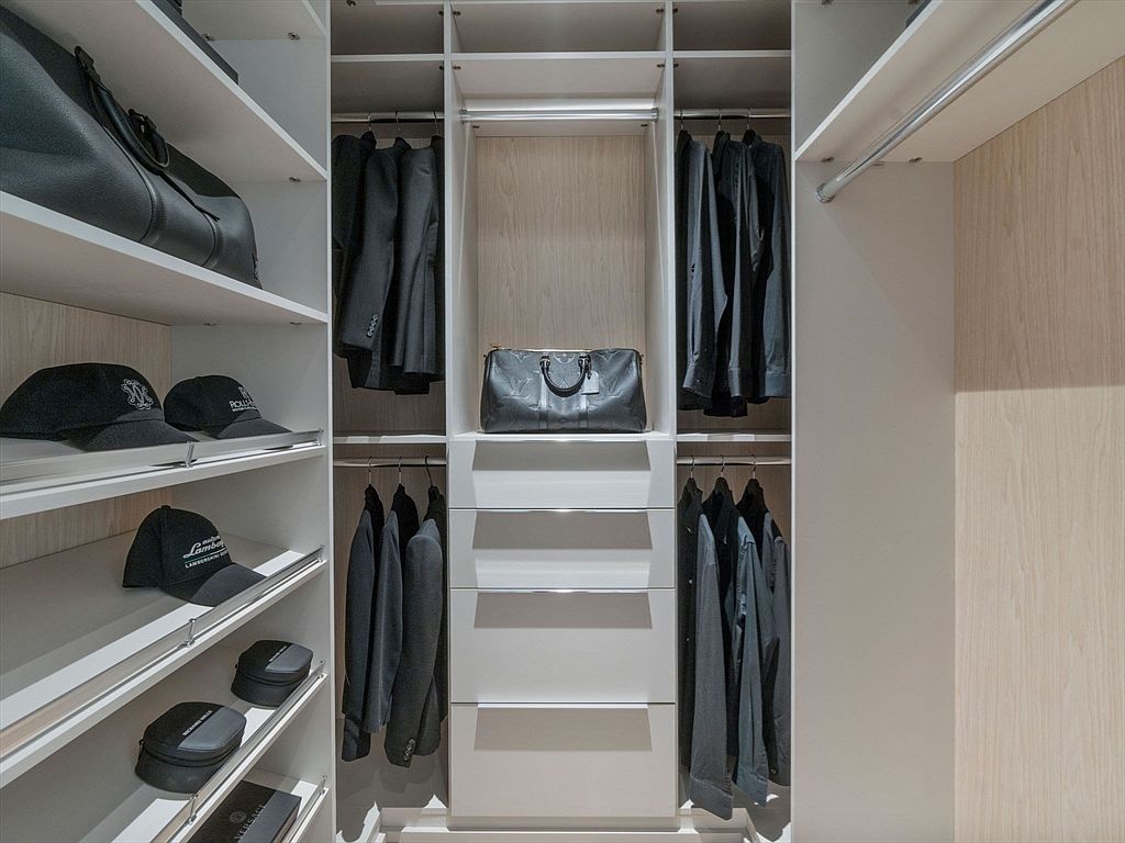 This is a well-organized walk-in closet featuring custom shelving and hanging rods. The closet is designed with a neutral color palette, including white shelves and light wood paneling. The space is efficiently used to store clothing, bags, and accessories, creating a tidy and luxurious feel.