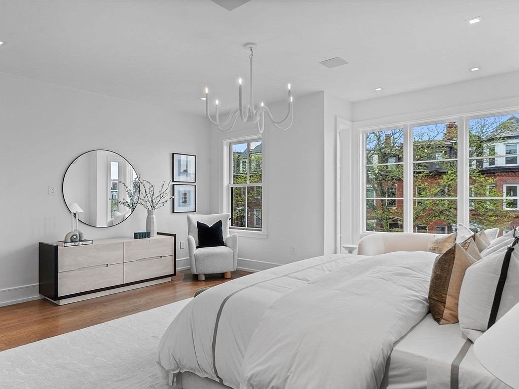 This is a bright and airy primary bedroom featuring a large bed with white linens and decorative pillows, a modern dresser with a round mirror above, and a comfortable armchair. Large windows provide ample natural light and views of the outside, while a stylish chandelier adds a touch of elegance. The room is decorated in a neutral color palette, creating a serene and inviting atmosphere.