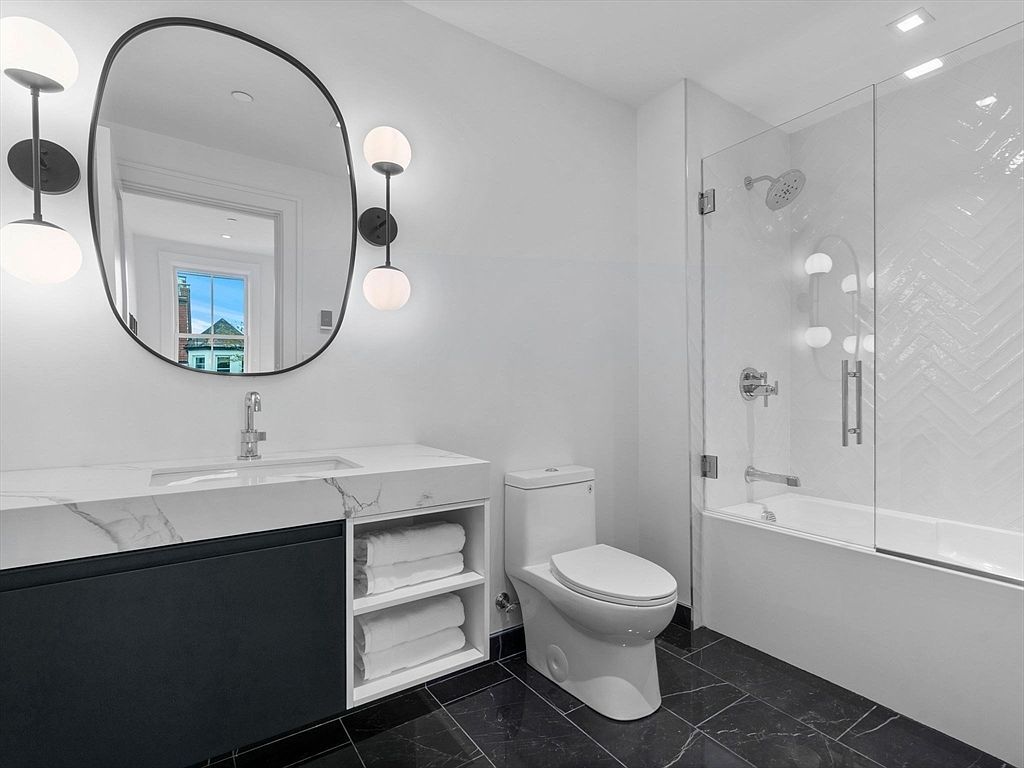 This is a modern bathroom featuring a black and white color scheme. The vanity has a marble countertop with a dark blue cabinet and open shelving for towels. A black-framed mirror hangs above the sink, flanked by sconce lighting, and a glass-enclosed shower/tub combination is visible to the right. The floor is tiled in a dark marble pattern, creating a sleek and contemporary look.