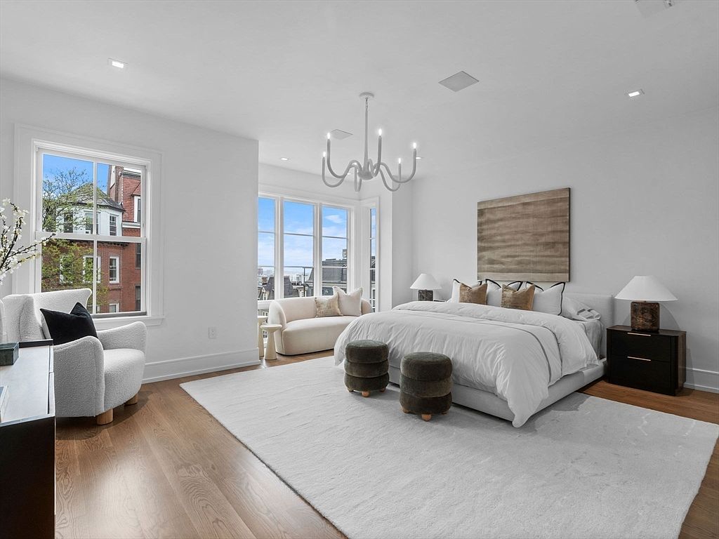 This is a bright and spacious primary bedroom featuring a large bed with white linens, flanked by dark wood nightstands and lamps. A seating area with a cream-colored sofa is positioned near a window offering city views. The room is decorated in a neutral color palette with a large area rug, creating a serene and luxurious atmosphere.