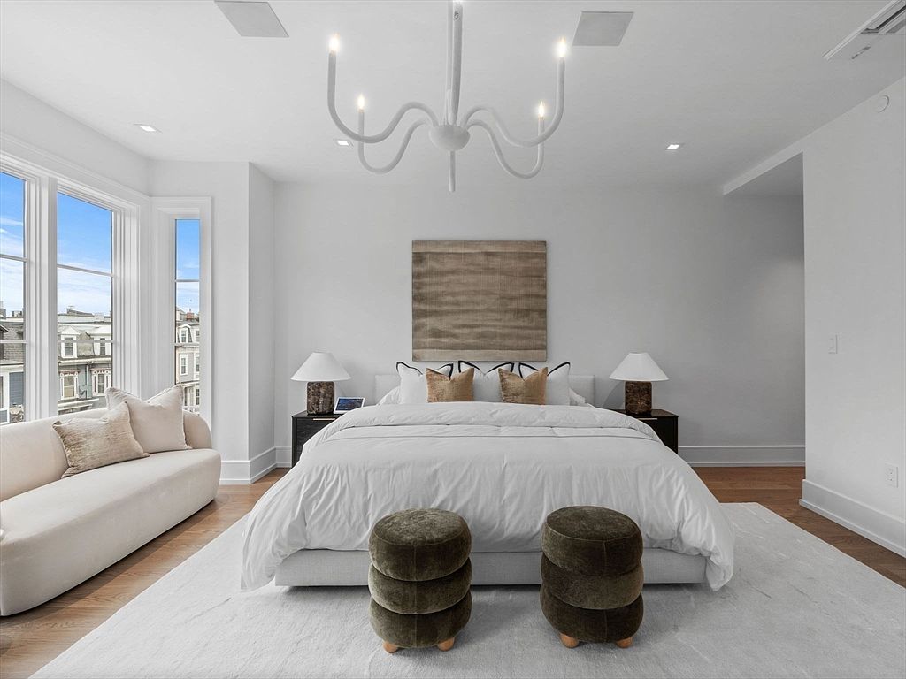 This is a bright and airy primary bedroom featuring a large bed with white linens, flanked by dark wood nightstands and lamps. A neutral-toned sofa sits near a window offering natural light and a view. The room is decorated in a minimalist style with a modern chandelier and a large piece of artwork above the bed, creating a serene and luxurious atmosphere.