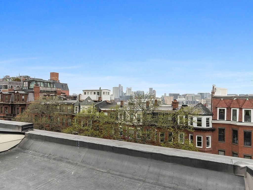 This aerial view showcases a cityscape with a focus on the rooftops of residential buildings. The buildings feature a mix of architectural styles, including brick facades and varying rooflines. In the distance, the city skyline is visible under a clear blue sky, creating a sense of urban living and proximity to city amenities.